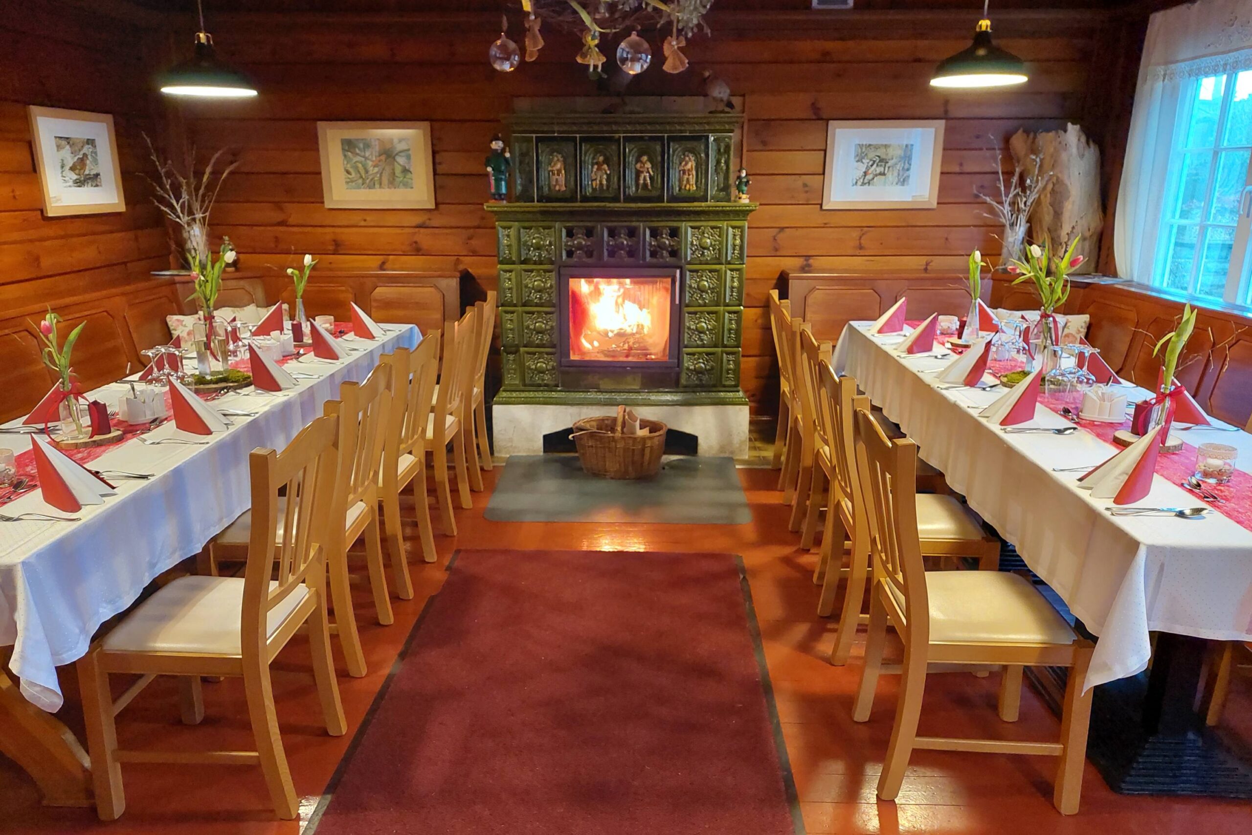 Das stimmungsvolle Kaminzimmer im Berggasthof Czorneboh bietet eine warme, einladende Atmosphäre. Zwei festlich gedeckte Tafeln mit weißen Tischdecken, roten Servietten und frischen Tulpen schaffen den perfekten Rahmen für Feiern und gesellige Abende. Im Mittelpunkt sorgt ein kunstvoll verzierter, grüner Kachelofen mit loderndem Feuer für wohlige Wärme. Holzvertäfelte Wände, liebevolle Dekorationen und große Fenster mit Blick ins Grüne runden das behagliche Ambiente ab.