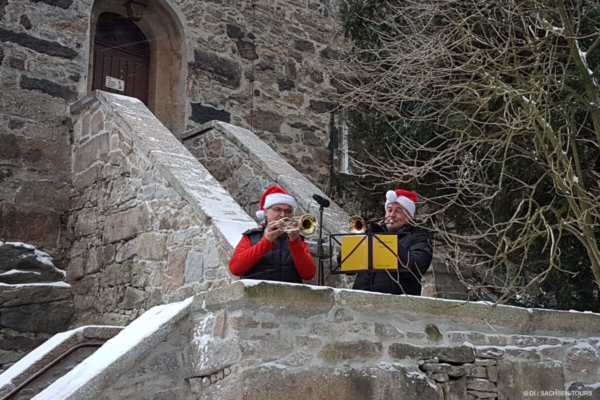 Zwei Personen mit Weihnachtsmannmützen spielen auf einer verschneiten Steintreppe vor dem historischen Czorneboh-Gebäude Turmblasen auf Trompeten. Einer von ihnen hält Notenblätter auf einem gelben Ständer, die die festliche Szene inmitten kahler Bäume und Schneeflecken noch verstärken.