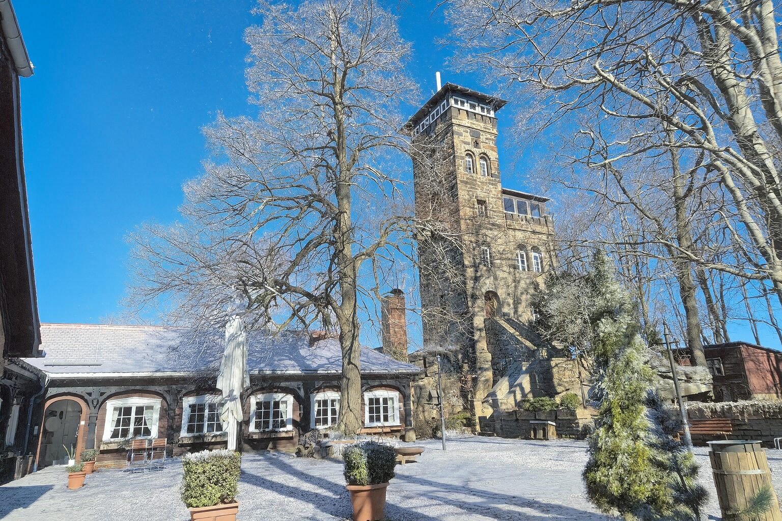 Hinter blattlosen Bäumen und einem Gebäude mit weiß gerahmten Fenstern erhebt sich an einem verschneiten Tag im Berggasthof Czorneboh ein Steinturm. Bänke, Tische und Topfpflanzen säumen den schneebedeckten Innenhof unter einem klaren blauen Himmel.