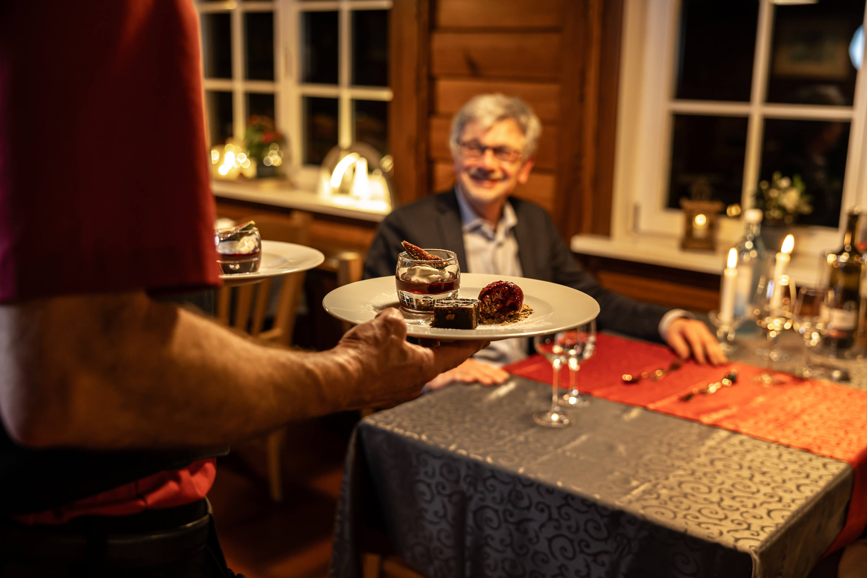 Ein Kellner serviert einem lächelnden Gast an einem kerzenbeleuchteten Tisch in einem gemütlichen Restaurant mit Holzwänden und warmer Beleuchtung Dessert und Getränke.